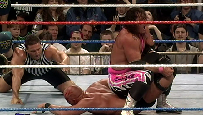 Bret Hart sits over Steve Austin's legs, holding him in the Sharpshooter submission as Austin lies on his stomach, grimacing in pain. Side profile picture. Referee Ken Shamrock looks on from the left.